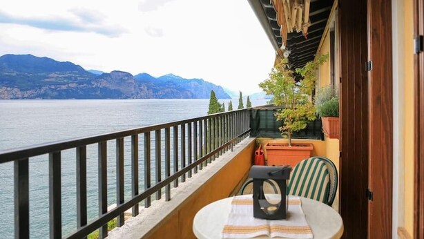 Un balcón con vistas a un lago y montañas, una mesa con una silla, y plantas en macetas.