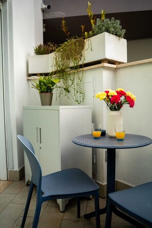 Um pequeno espaço ao ar livre com uma mesa redonda, duas cadeiras, um armário com plantas e um vaso com flores.