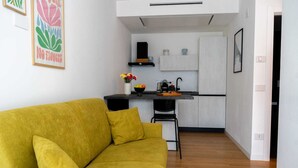 Un espacio habitable moderno con un sofá amarillo, una cocina con armarios blancos y una silla negra.