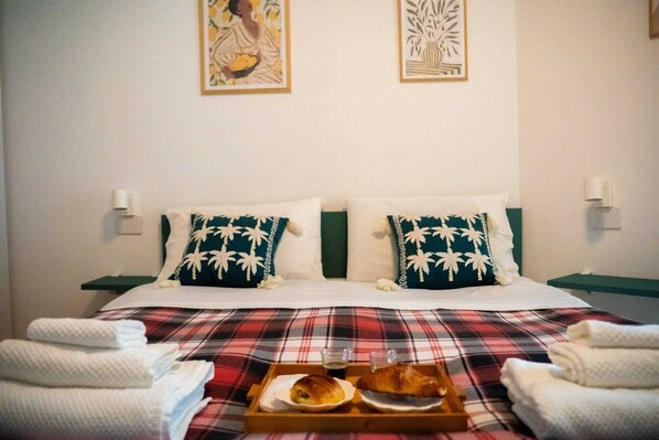 Uma cama de casal com colcha xadrez, duas almofadas com desenhos de folhas de palmeira, um bandeja com croissants e café, e duas mesas de cabeceira com abajures.