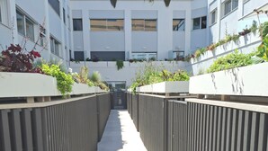 Un sendero estrecho bordeado por paneles de madera verticales con follaje en la parte superior, que conduce a la entrada de un edificio.
