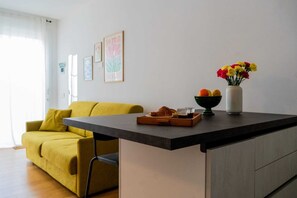 Uma sala de estar moderna com um sofá amarelo, uma pequena ilha de cozinha com um tabuleiro de comida e bebidas, e um vaso de flores.
