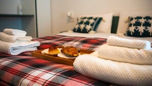 Una cama con una manta escocesa, almohadas, una bandeja con croissants y un vaso de jugo, y toallas dobladas.