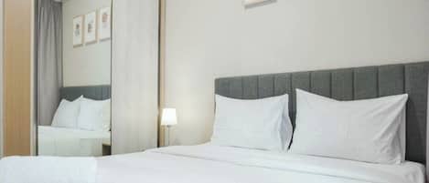 Una habitación de hotel con una cama grande, cabecera gris, ropa de cama blanca, una lámpara de noche y cuadros en la pared.