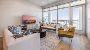 A modern living room with a sofa, armchair, and coffee table, large windows with a city view, and a flat-screen TV on the wall. - @ Marbella Lane - Panoramic Delight Dream Escape! (Long Beach)