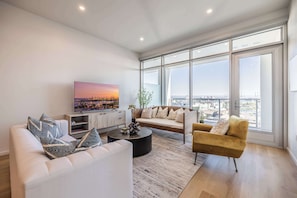 A modern living room with a sofa, armchair, and coffee table, large windows with a city view, and a flat-screen TV on the wall. - @ Marbella Lane - Panoramic Delight Dream Escape! (Long Beach)