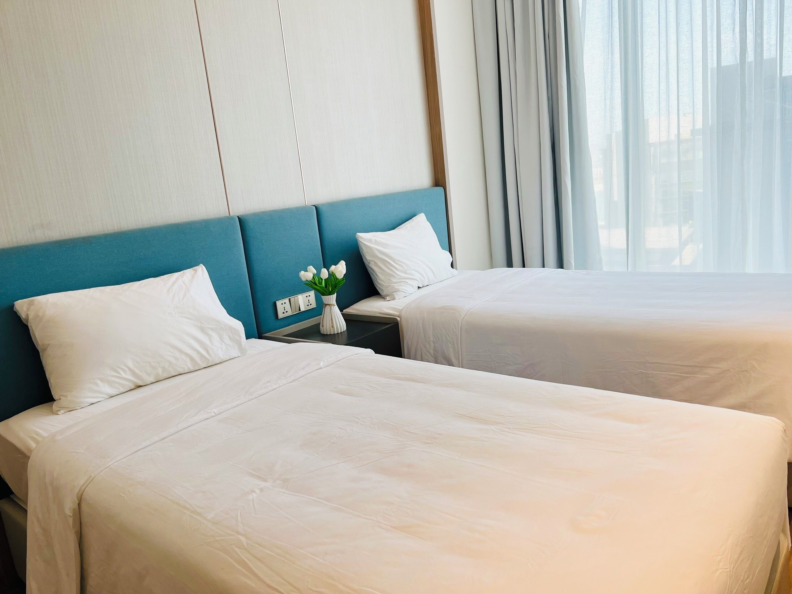 Two beds with white linens, a vase with flowers, and a power outlet on the wall.