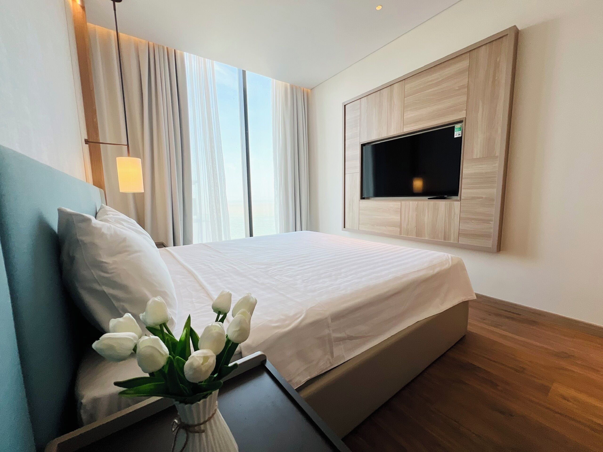 A modern hotel room with a bed, a flat-screen TV mounted on the wall, a bedside table, and a window with curtains.