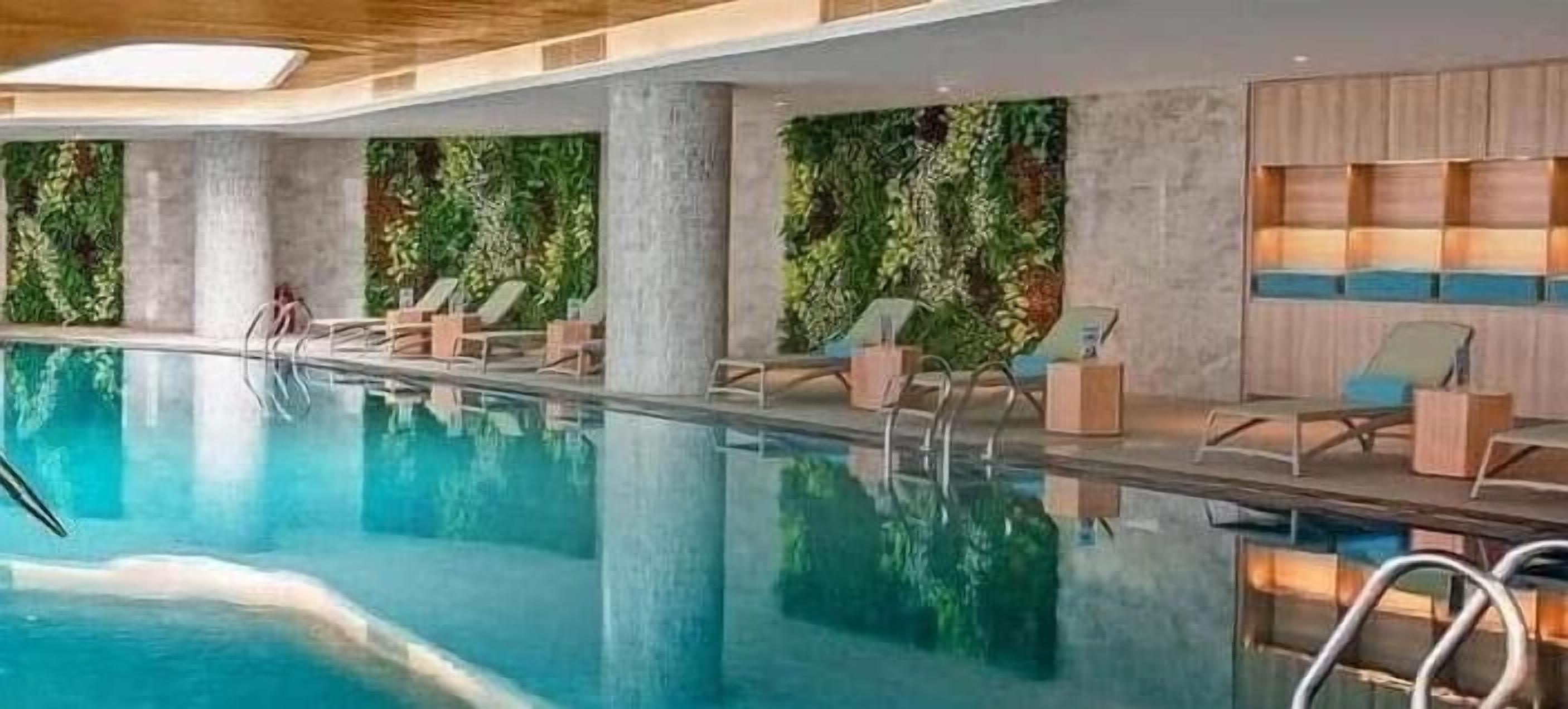 A modern indoor swimming pool with clear water, surrounded by lounge chairs and a wall with vertical greenery.