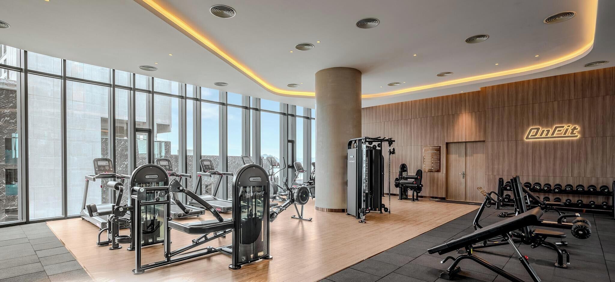 A modern gym with various exercise machines, large windows, and a wooden wall with a neon sign.