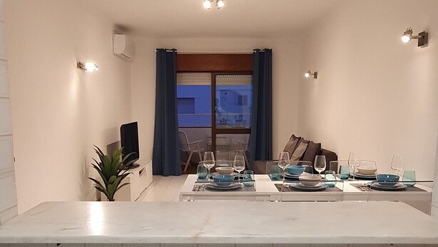 A modern kitchen with a marble countertop, a dining area with a table set for four, and a view of a blue building through a window.