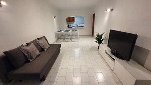 A living room with a sofa, a TV, a small kitchen area, and a tiled floor.