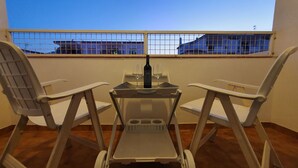 A balcony with two white plastic chairs, a small table with a bottle and two glasses, and a view of buildings through a metal fence.