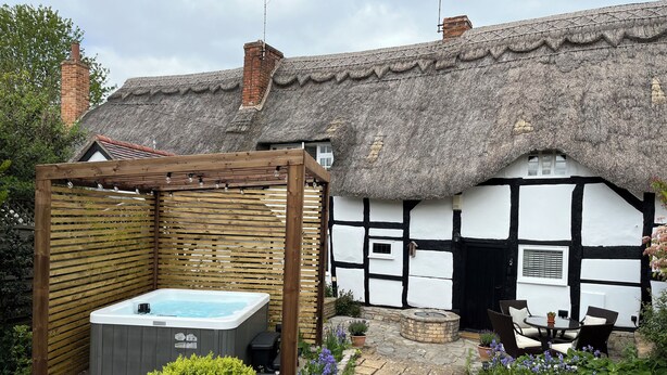 A traditional thatched-roof house with a wooden pergola and a hot tub in the garden.