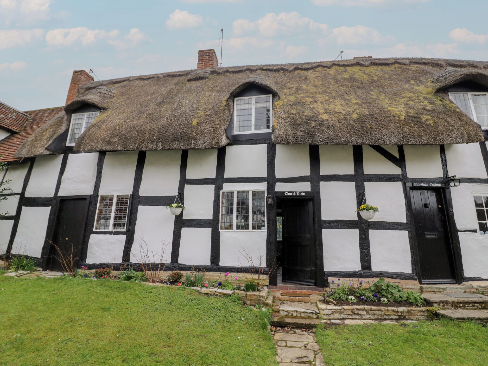 A traditional Tudor-style building with a thatched roof, black and white timber framing, and a small garden area with stepping stones leading to the entrance.