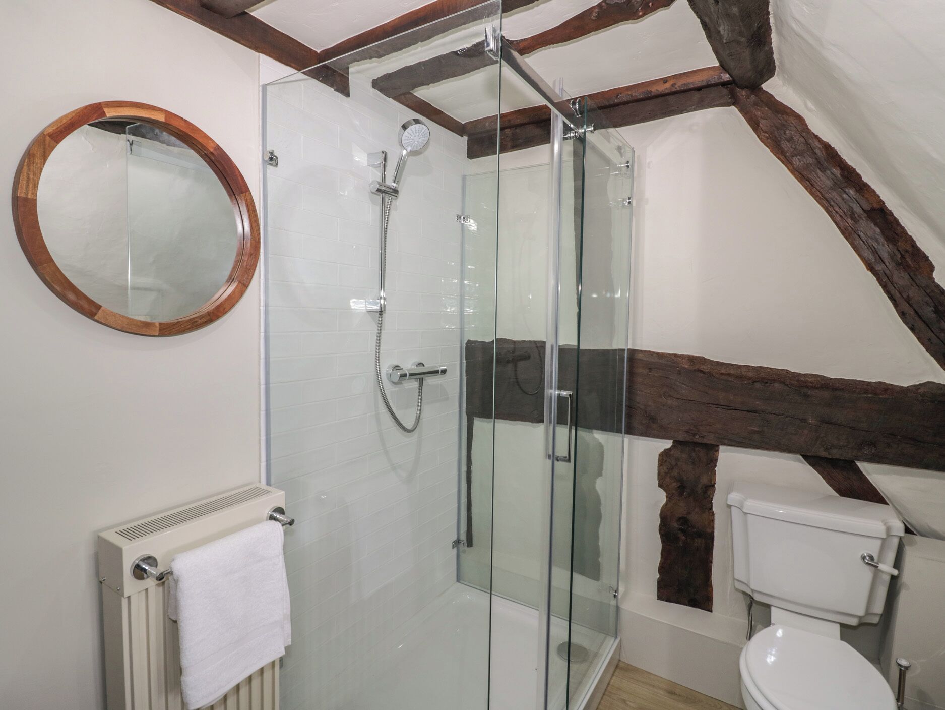 A modern bathroom with a glass-enclosed shower, a round mirror, a toilet, and a radiator.