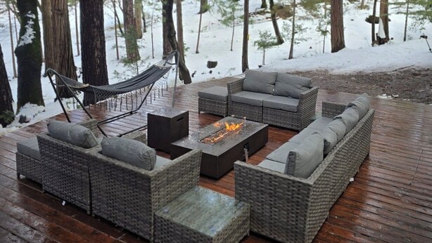 Une terrasse en bois, aménagée avec du mobilier d’extérieur, notamment des canapés et un foyer, le tout entouré d’arbres.