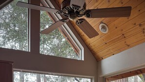 Une cuisine avec des meubles de rangement en bois, un ventilateur de plafond et de grandes fenêtres donnant sur la verdure.