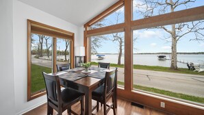 Un espace repas comprenant une table et des chaises, de grandes fenêtres donnant sur un lac et un quai, et un plancher en bois.