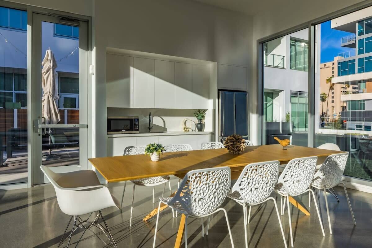 A modern dining area with a wooden table and white chairs, large windows offering views of buildings, and a kitchen area with cabinets and appliances.