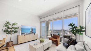 A modern living room with a large TV, a sofa, a coffee table, and a view of the city through large windows.