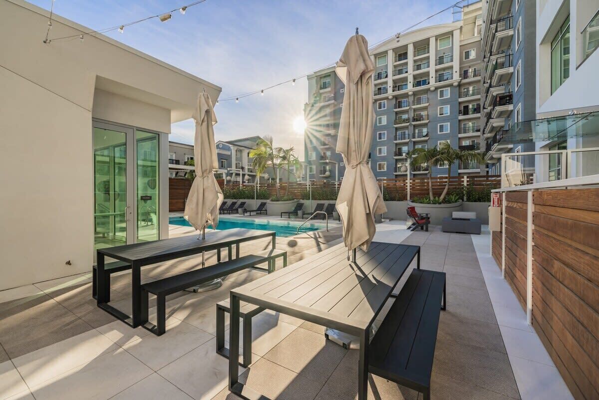 A rooftop pool area with lounge chairs and umbrellas, a wooden deck, and a view of a high-rise building in the background.