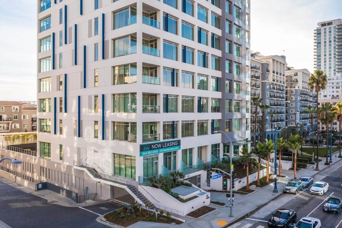 A modern multi-story building with a prominent 'NOW LEASING' sign, featuring balconies, large windows, and a parking area with cars.