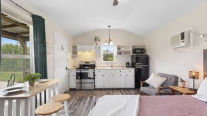 A compact kitchen with a microwave, stove, and sink, adjacent to a bedroom with a bed and a nightstand.