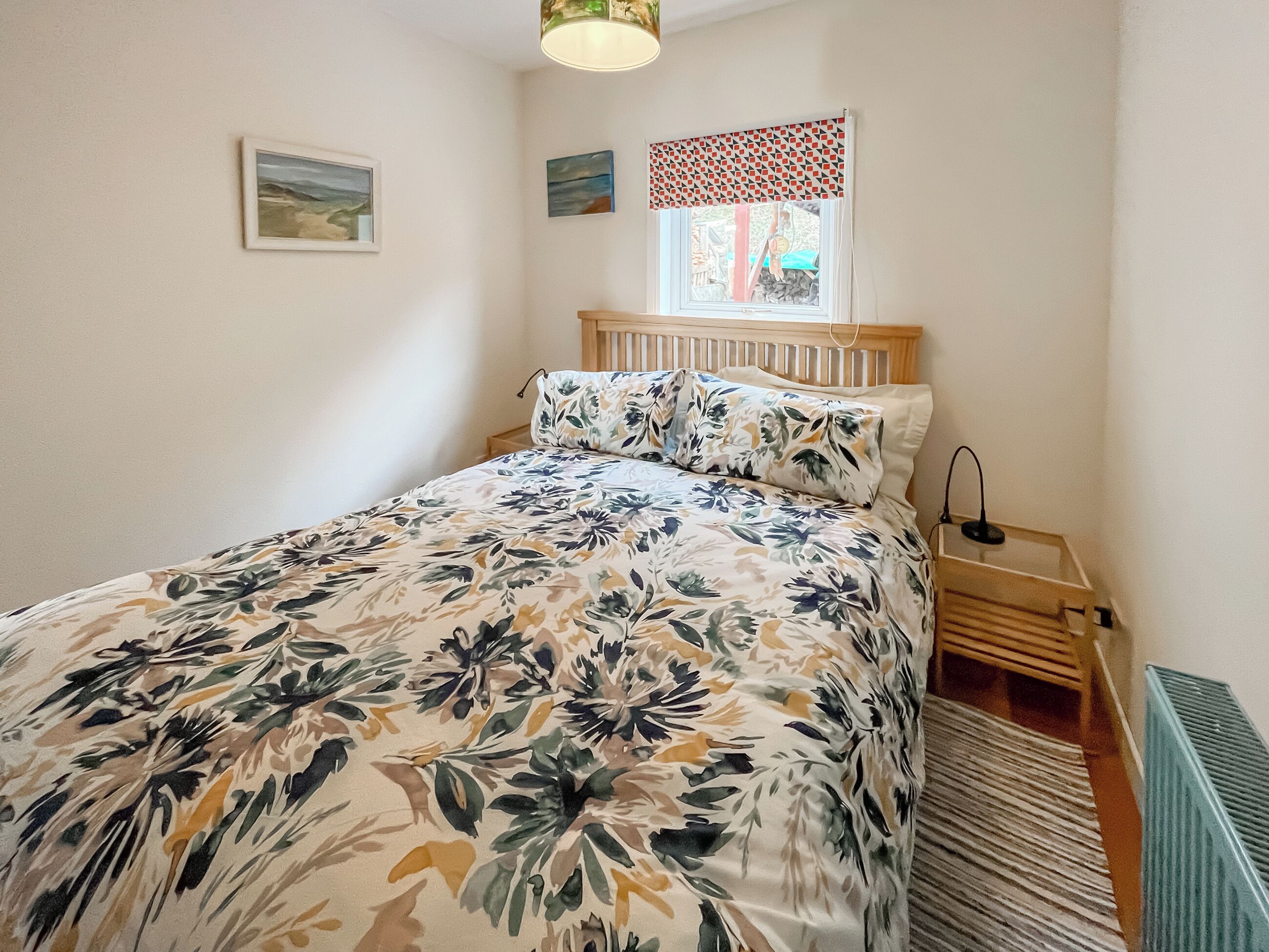 Un dormitorio con colcha estampada, cabecera de madera, mesita de noche con lámpara y ventana con cortinas decoradas.