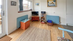 A living room with a wooden floor, a blue radiator, a television, a bookshelf, and a painting on the wall. - Ardtornish Apartment (Tayvallich)
