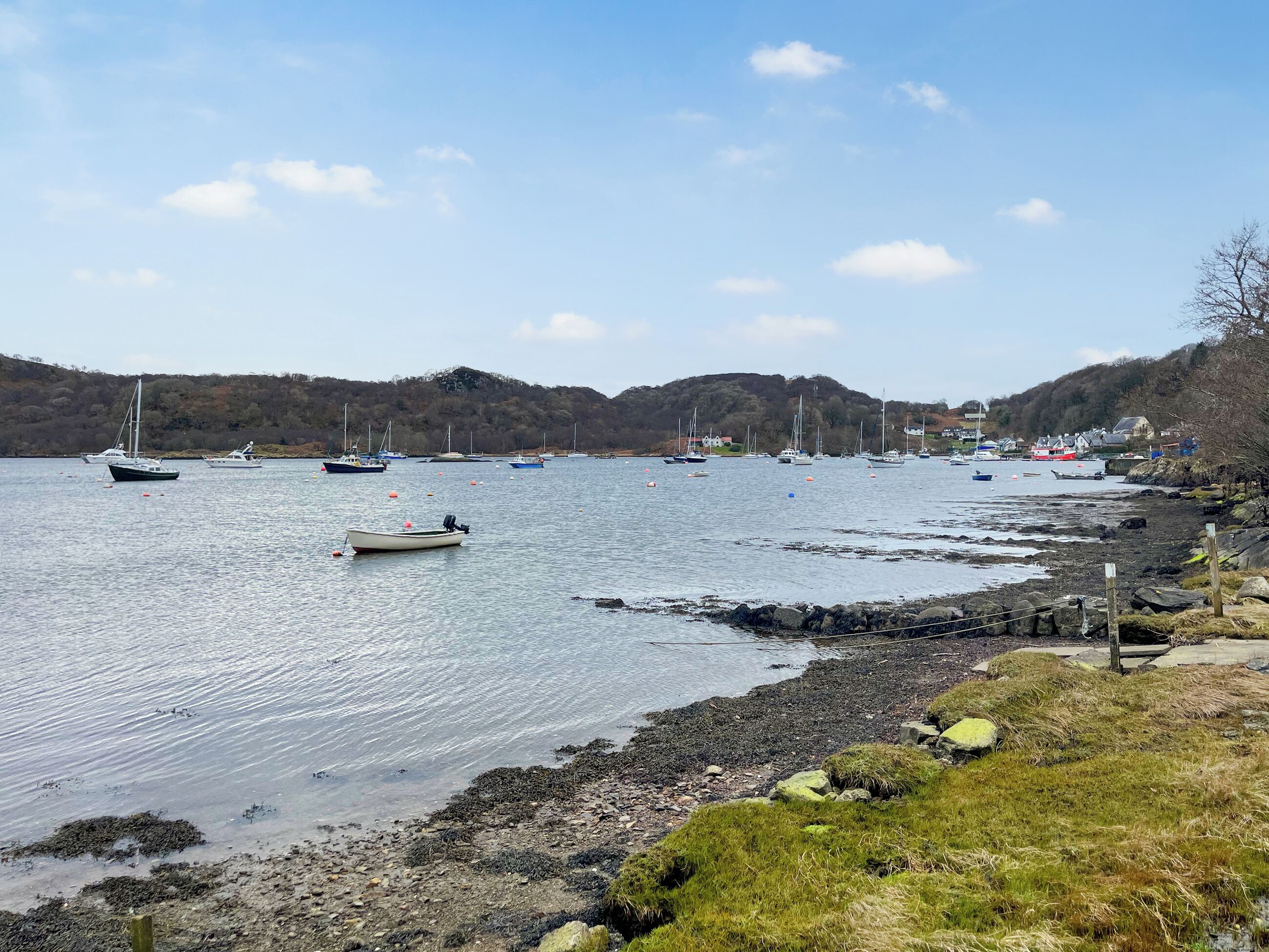 A coastal scene with boats on calm water, rocky shoreline, and hills in the background.