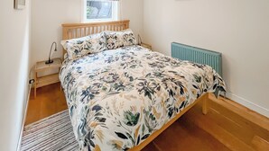 A bedroom with a wooden bed, a bedside table with a lamp, a radiator, and a window with a patterned blind. - Ardtornish Apartment (Tayvallich)