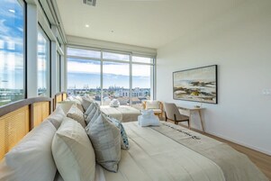 Habitación de hotel con un ventanal grande, una cama con colcha blanca, una silla, una mesita y un cuadro en la pared.