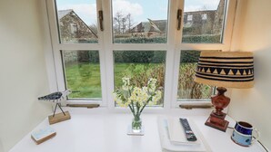 Ein Fenster mit Blick auf einen Garten und Häuser, ein Schreibtisch mit Blumen in einer Vase, eine Fernbedienung und eine Lampe.