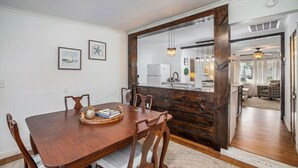 Comedor con mesa y sillas de madera, cocina visible a través de un panel de vidrio y sala de estar con sofá y cortinas.