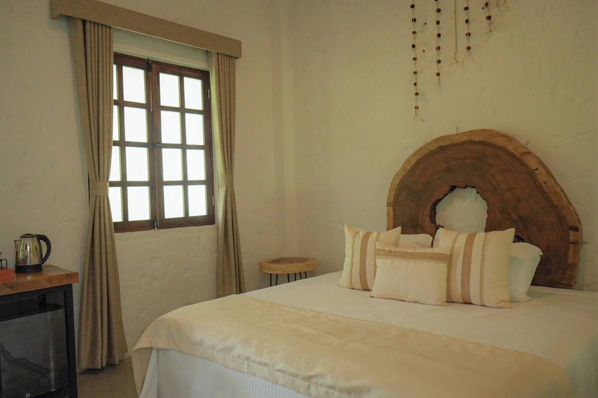 A bedroom with a large bed, a wooden headboard, a window with curtains, and a small table with a kettle.