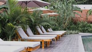 A row of wooden lounge chairs with white cushions, arranged along a tiled poolside area with large umbrellas providing shade. - Anzalik Hotel Boutique (Anza)