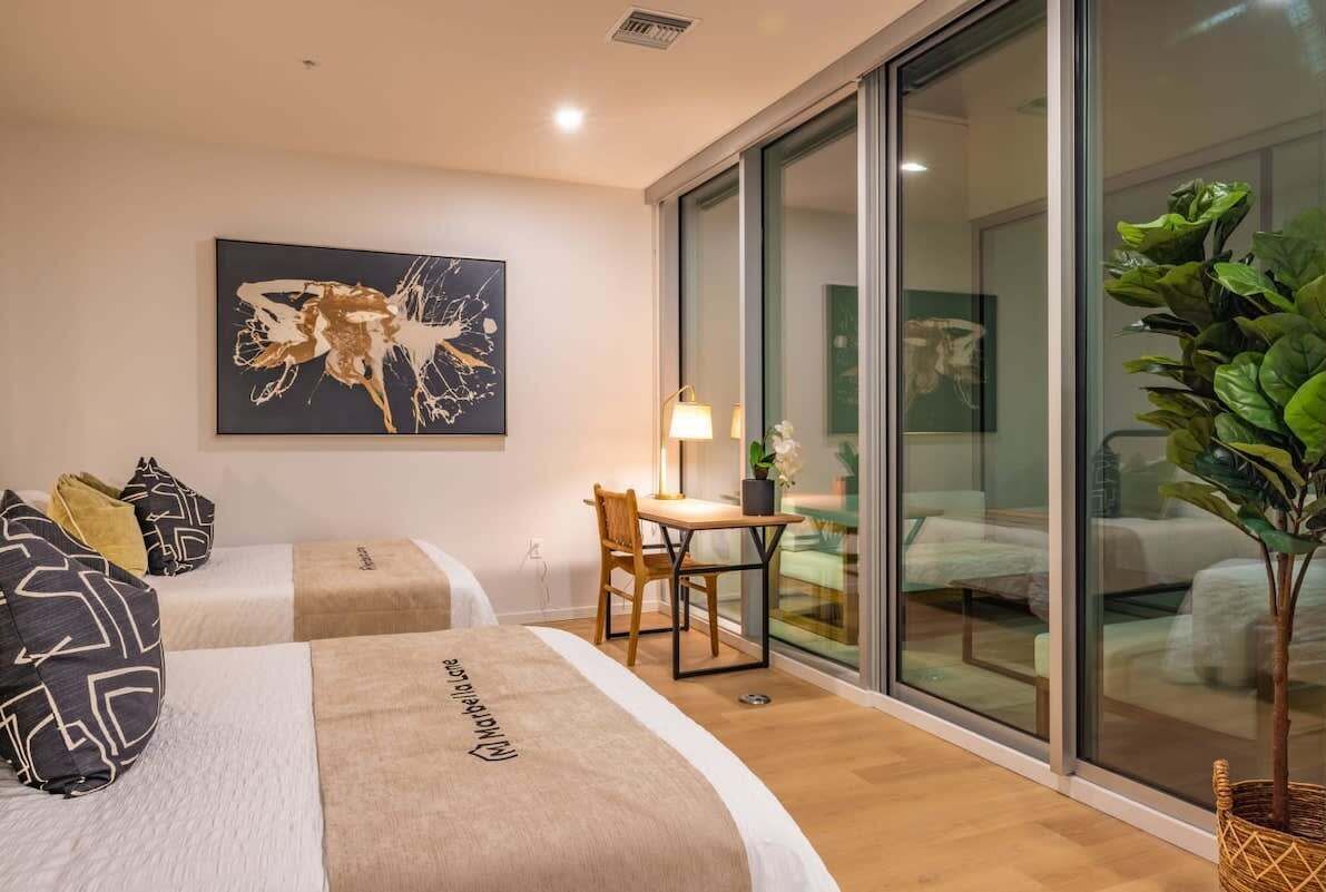 A hotel room with two beds, a desk, a chair, a large window, and a plant.