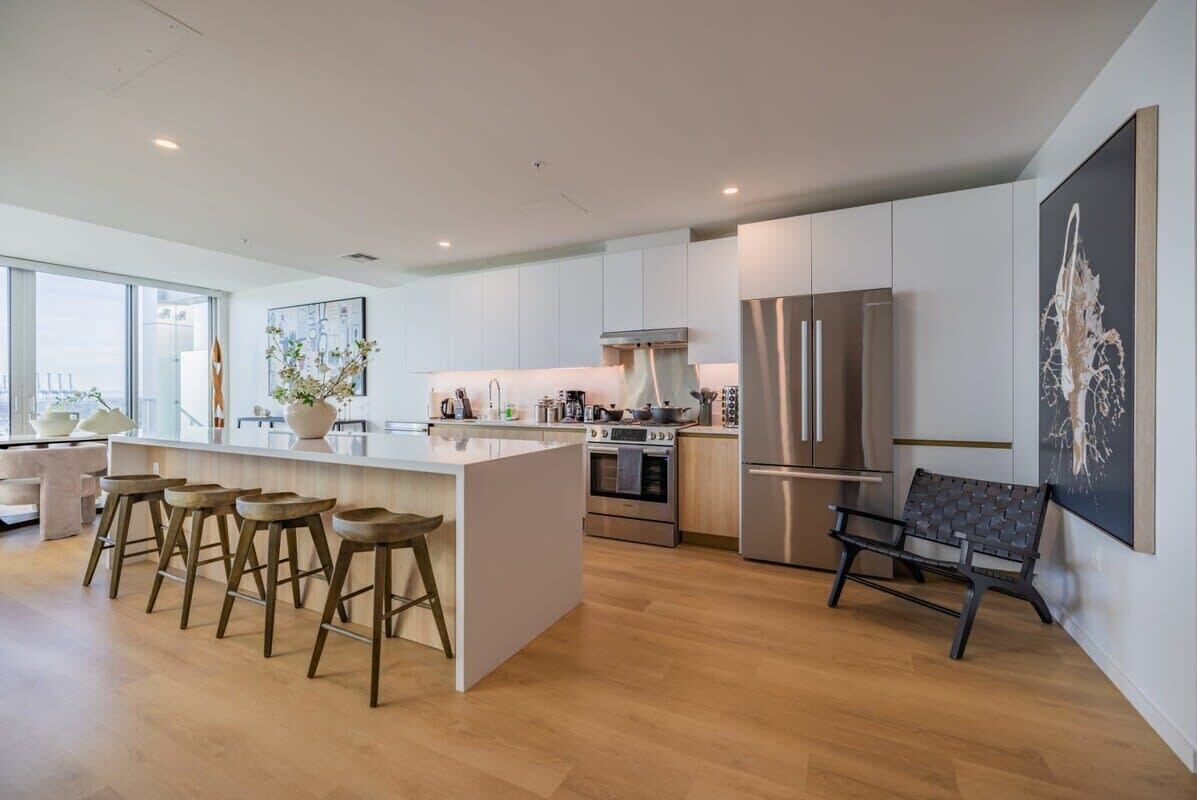 A modern kitchen with a central island, bar stools, stainless steel appliances, and a large window providing natural light.