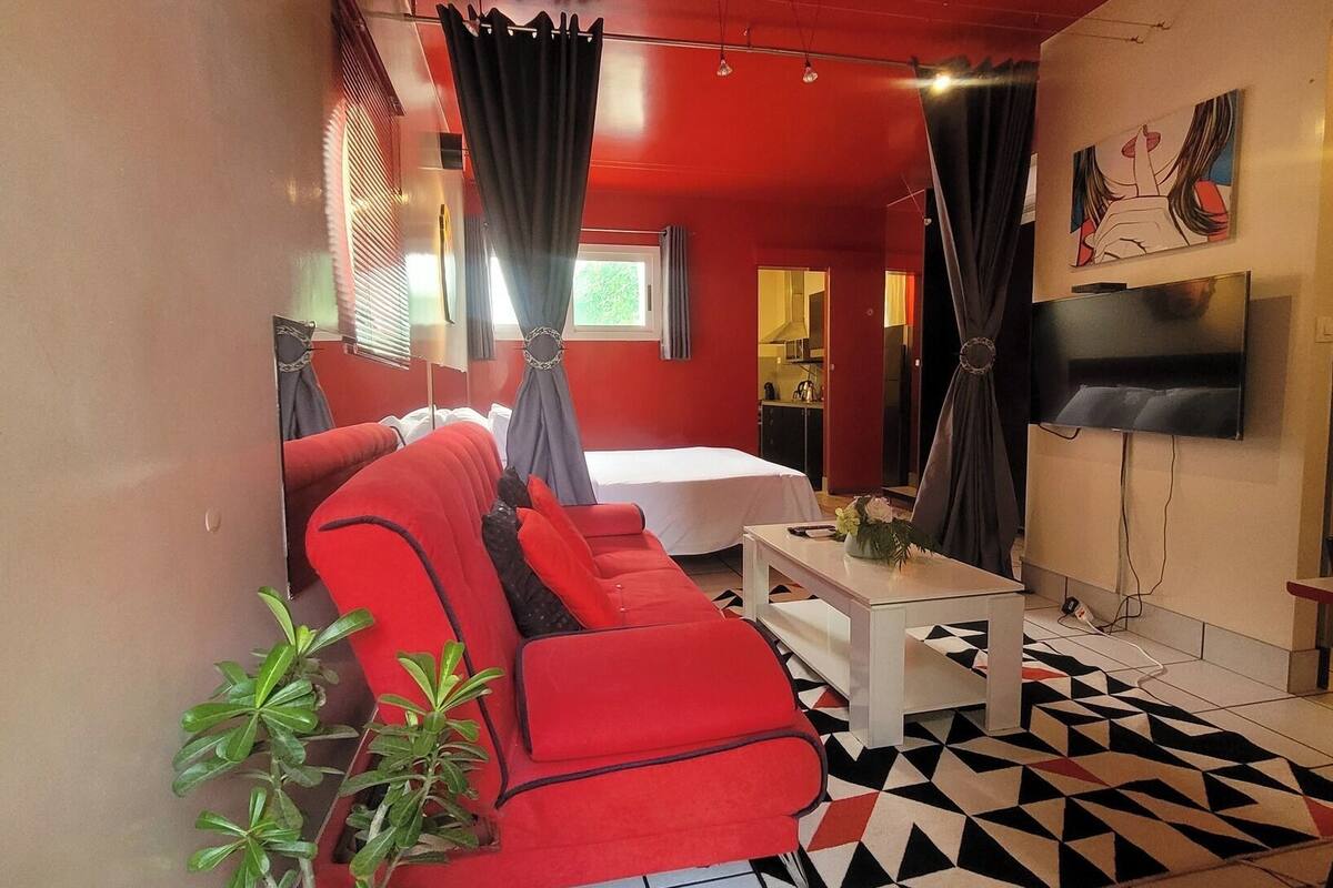 Une chambre d’hôtel de taille modeste, avec un canapé rouge, une petite table blanche, un téléviseur et un sol orné d’un motif géométrique.