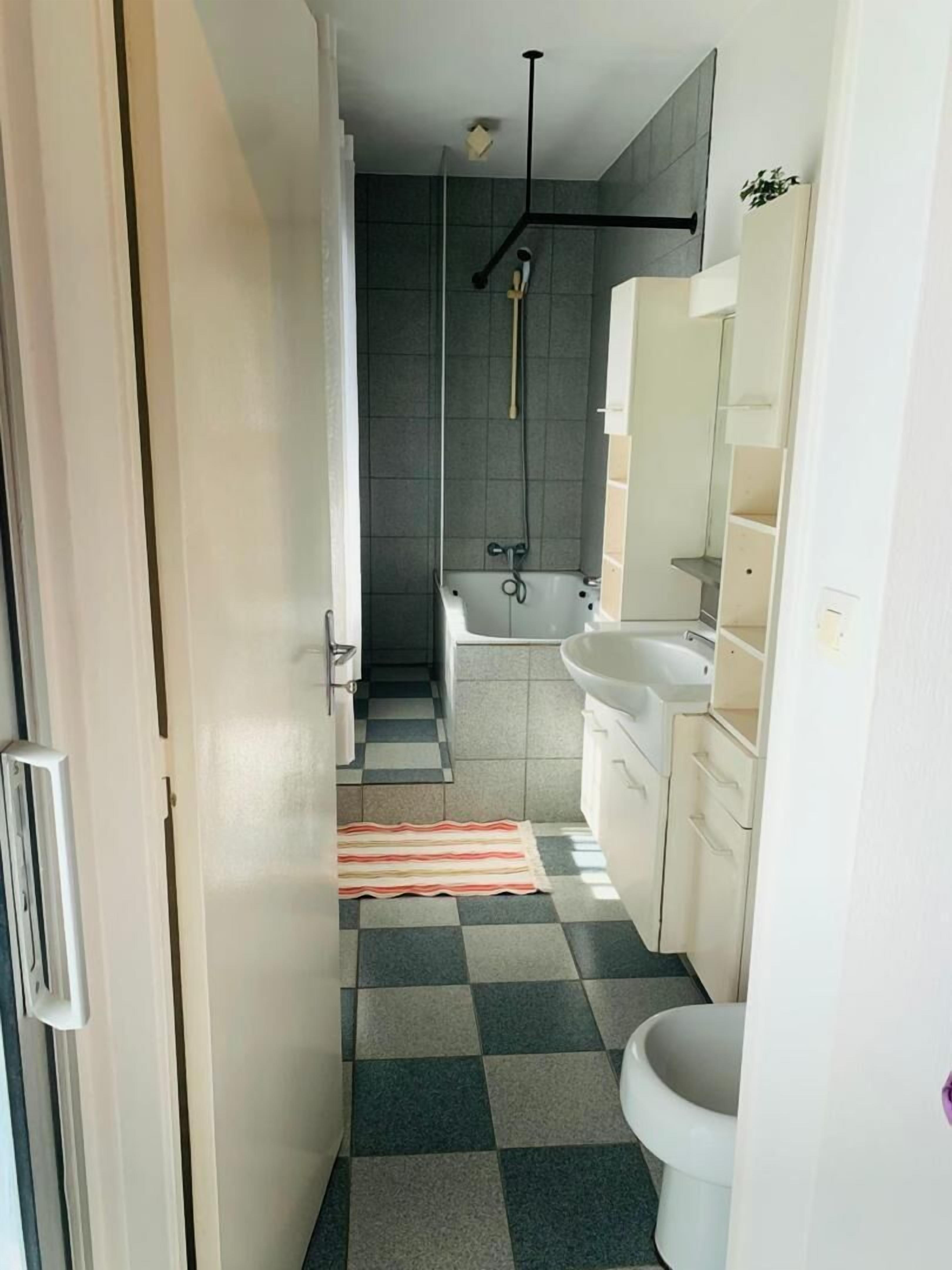 A narrow bathroom with a checkered floor, a bathtub, a sink, and a shower.