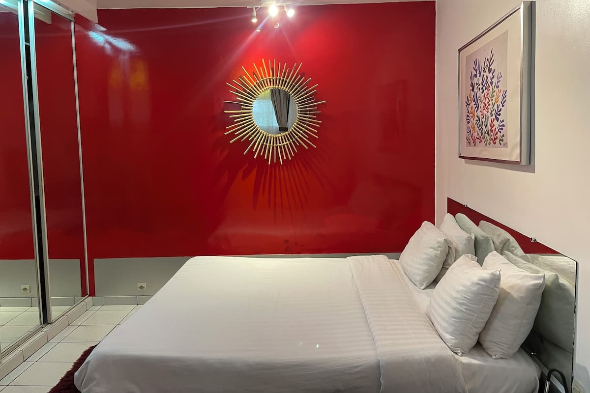 Une chambre à coucher moderne avec un grand lit, un mur d’accent rouge, un miroir original en forme de soleil et une œuvre d’art encadrée.