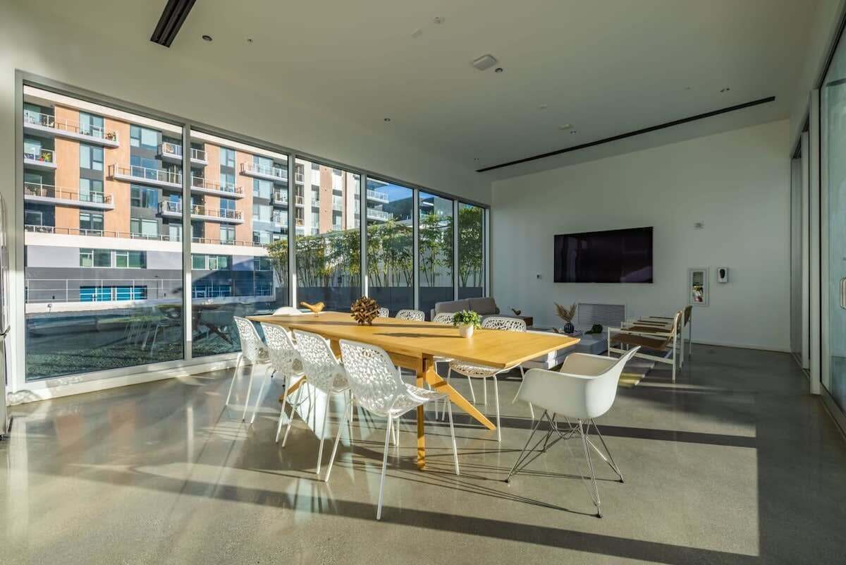 A modern conference room with a large table, white chairs, and large windows offering a view of a residential building.