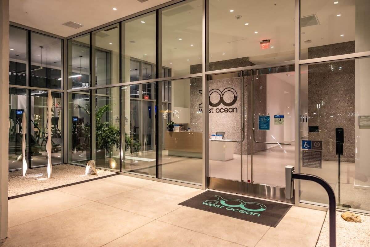 A modern hotel entrance with glass doors, a reception desk, and a digital display.