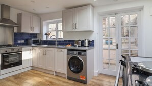 A modern kitchen with white cabinets, a built-in oven, a microwave, a sink, and a washing machine. There is a skylight above the sink and a view of the outside through large windows. - Starfish House (Lyme Regis)
