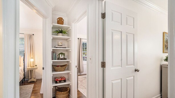 A hallway with a white door, a shelf unit with baskets, and a small table with a lamp.