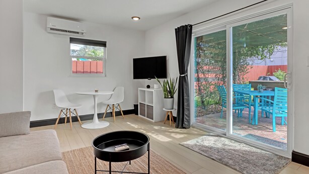 A modern living room with a sofa, a coffee table, a TV, and a sliding glass door leading to an outdoor patio with blue chairs and a grill. - Steps to Cocoa Beach, MIL Suite & Hot Tub, Amenities Galore!! (Cocoa Beach)