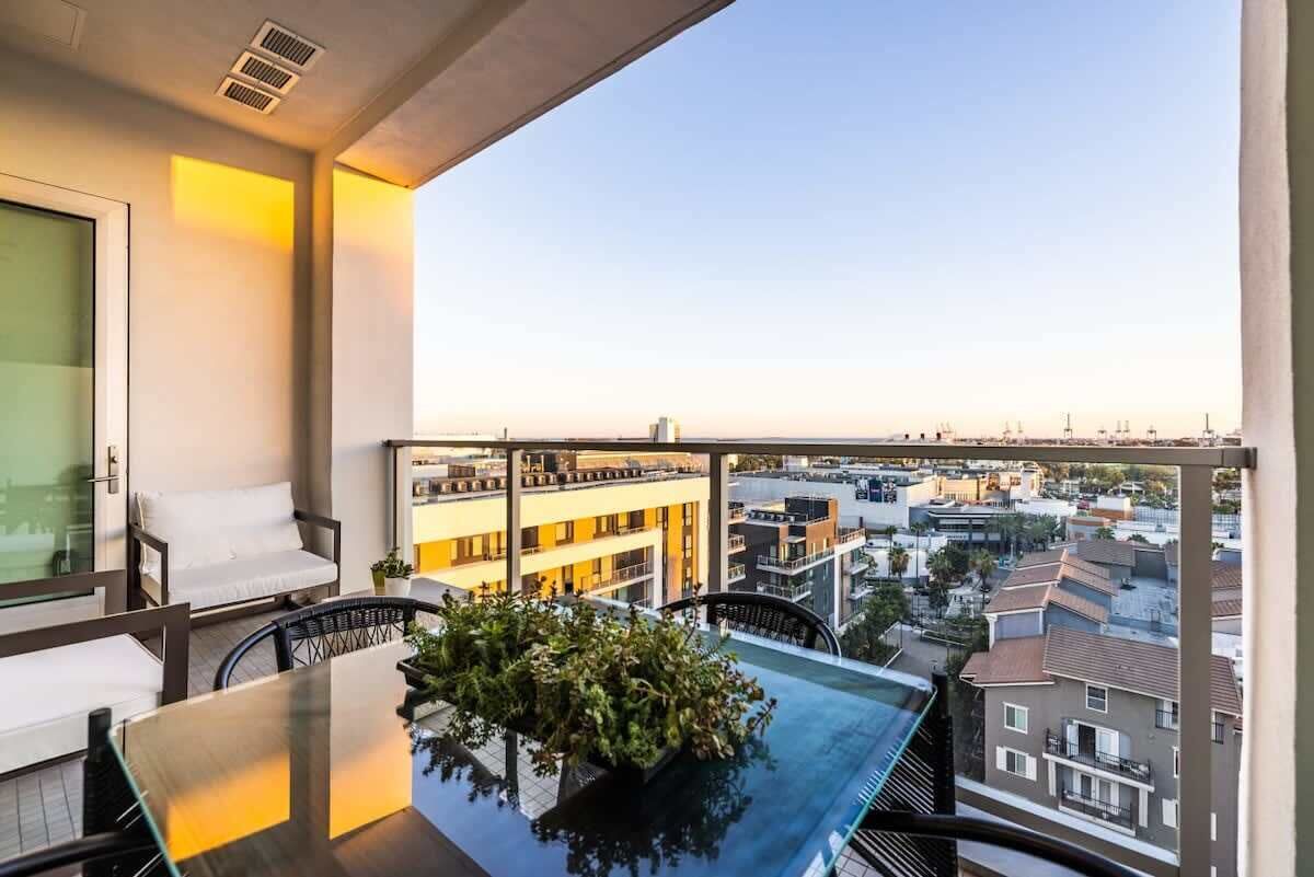 Een balkon met een glazen tafel, witte stoelen met kussens en uitzicht op de stad.