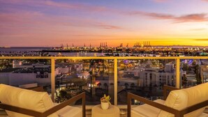 Ein Balkon mit zwei weißen, gepolsterten Stühlen und einem kleinen Tisch mit einer Pflanze, mit Blick auf eine Stadtlandschaft bei Sonnenuntergang.