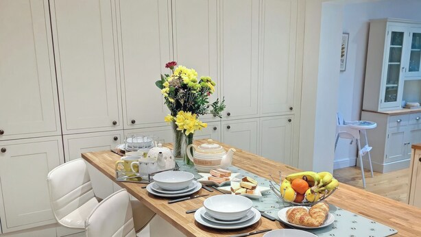 Un coin repas avec une table en bois dressée pour quatre personnes, des chaises blanches, un vase de fleurs jaunes et, au fond, une cuisine.